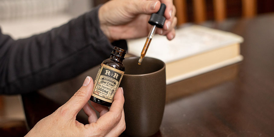 close up view of a person's hands holding a bottle of hemp extract and a dropper with cbd oil over a mug on top of a table